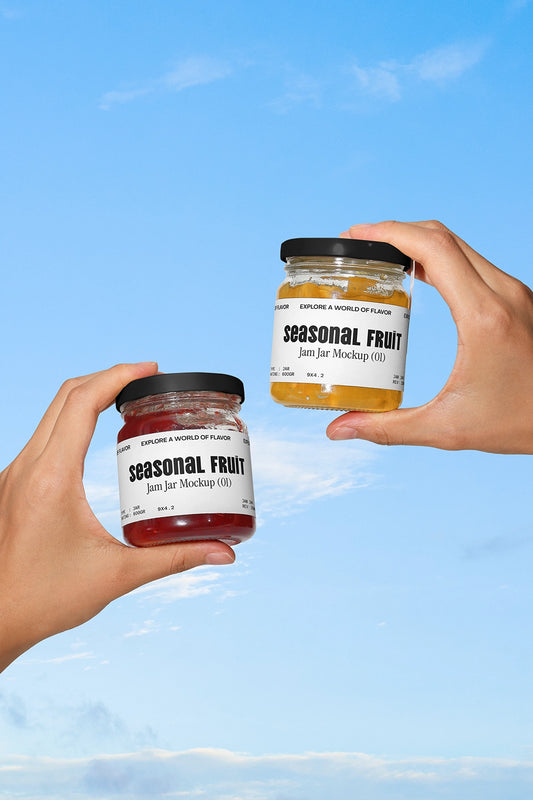 Hands holding two jam jars mockup with seasonal fruit labels against blue sky background