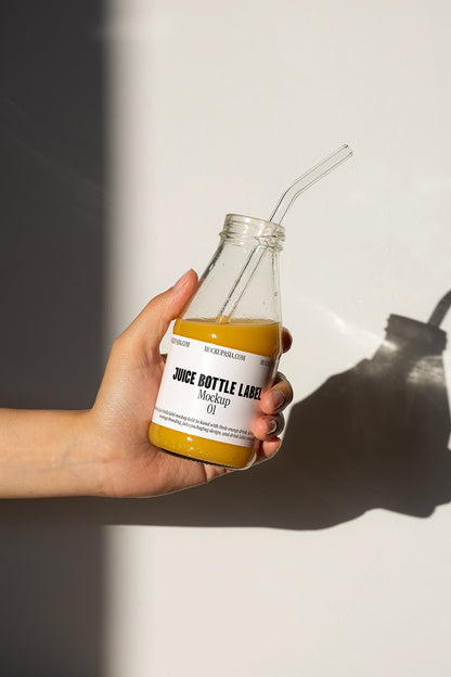 Hand holding glass juice bottle mockup with straw and orange juice in natural sunlight