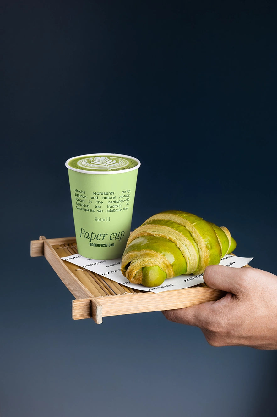 Hand-held cafe serving tray with matcha latte paper cup and green croissant against a dark blue background