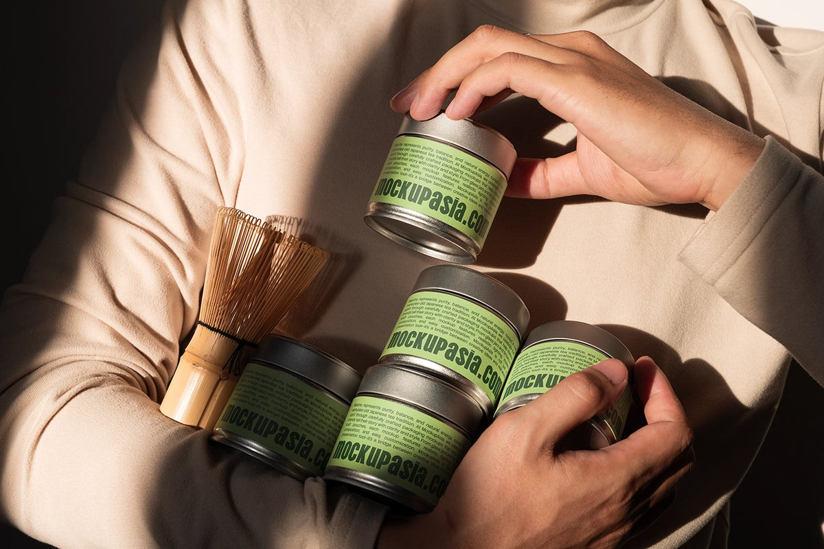 A person in a beige sweater holds several small silver tins with light green labels reading mockupasia.co, alongside a wooden whisk, in warm directional sunlight.