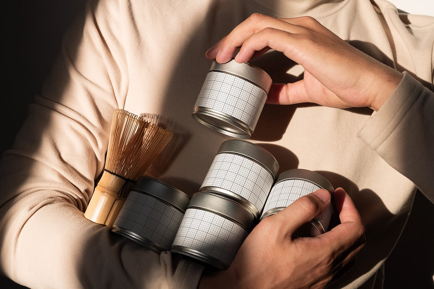 A person in a beige long-sleeve top holds several small silver metal tins with white grid-patterned labels, alongside a bamboo whisk, in warm directional sunlight.