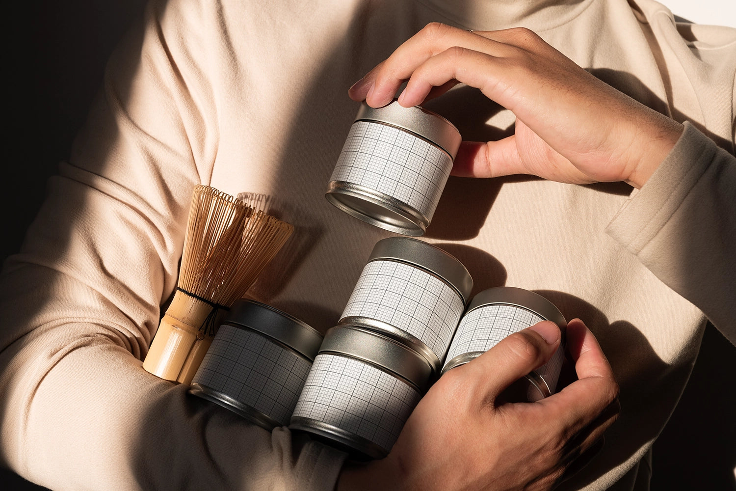 A person in a beige long-sleeve top holds several small silver metal tins with white grid-patterned labels, alongside a bamboo whisk, in warm directional sunlight.