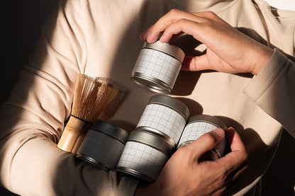A person in a beige long-sleeve top holds several small silver metal tins with white grid-patterned labels, alongside a bamboo whisk, in warm directional sunlight.