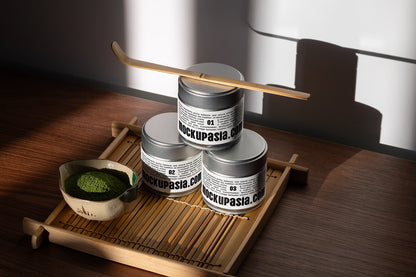 Realistic matcha tin can mockup with three metal tins on a bamboo tray beside matcha powder and a bamboo whisk