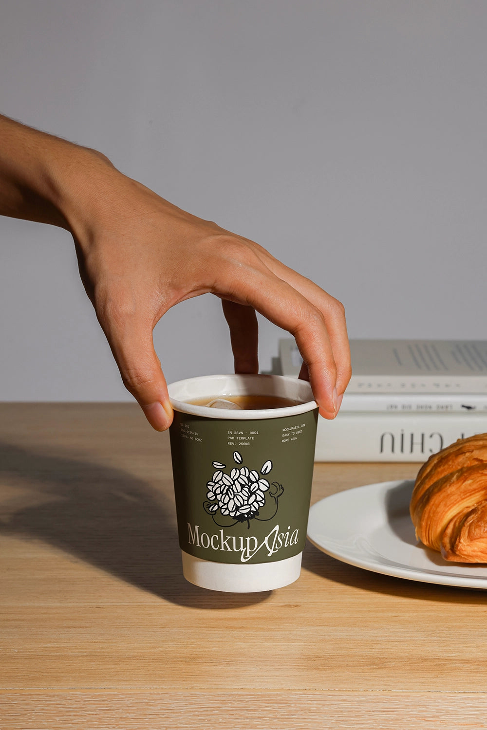 Hand holding a coffee cup mockup on a wooden table with a croissant and books in the background.