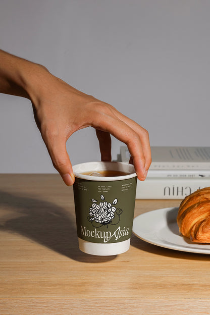 Hand holding a coffee cup mockup on a wooden table with a croissant and books in the background.