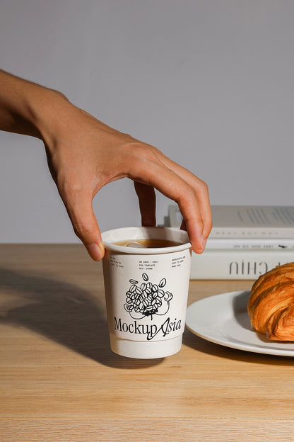 Hand holding a coffee cup mockup on a wooden table with a croissant and books.