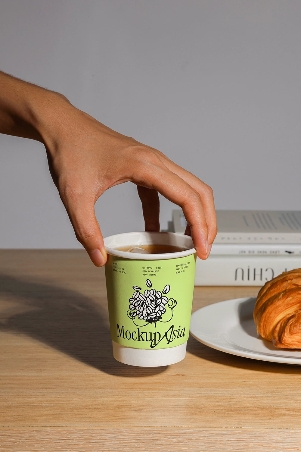 Hand holding a green coffee cup mockup psd with a croissant on a plate in the background