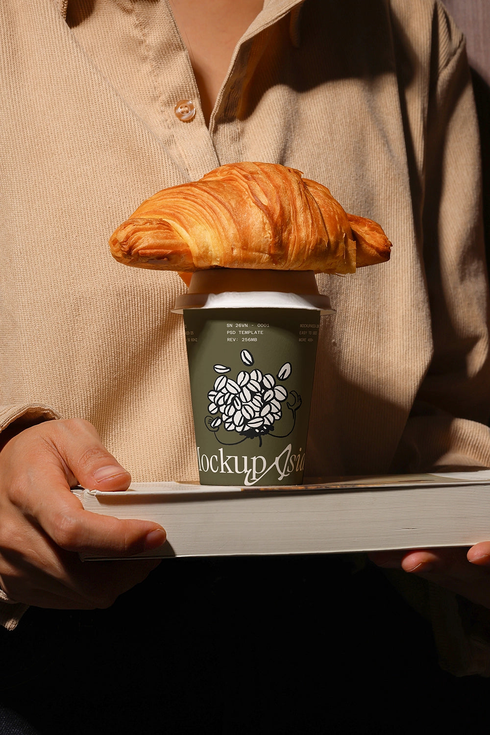 Person holding a coffee cup mockup with a croissant on top psd template