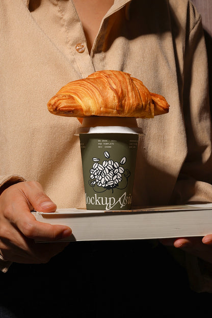 Person holding a coffee cup mockup with a croissant on top psd template