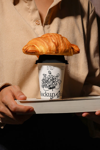 Person holding a coffee cup mockup with a croissant on top