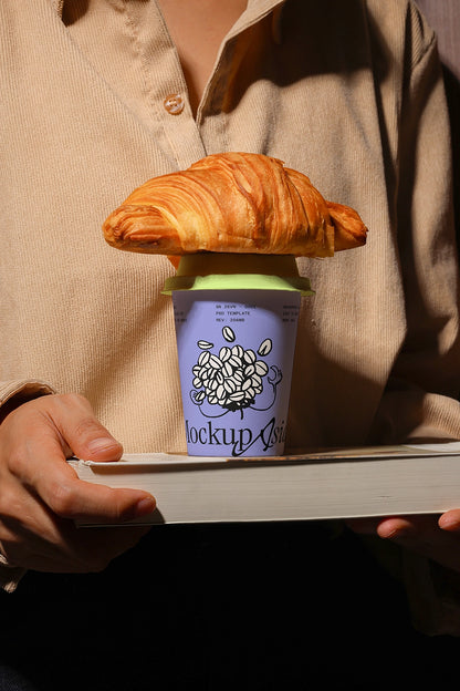Person holding a coffee cup with a croissant on top, brand 'mockup' visible