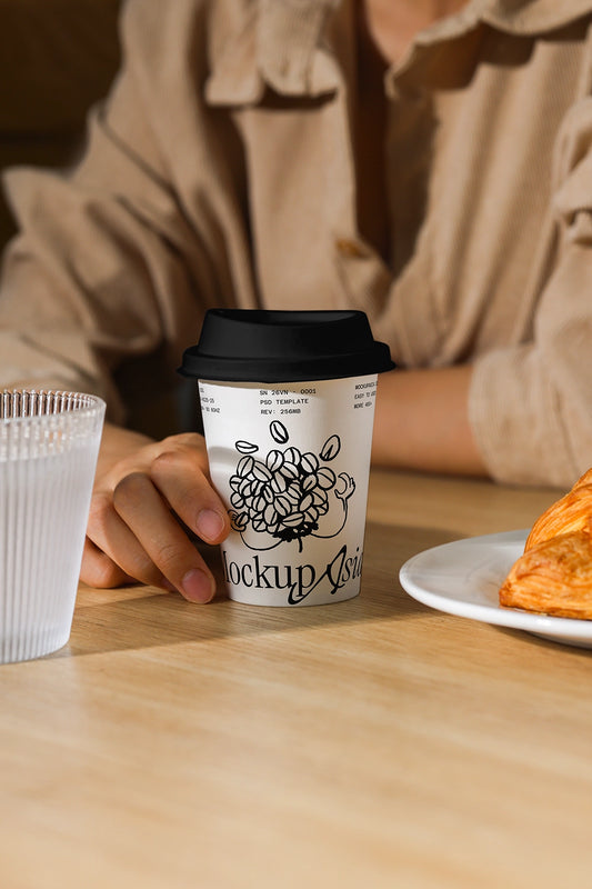 Person holding a coffee cup with a mockup design on a wooden table with a pastry.