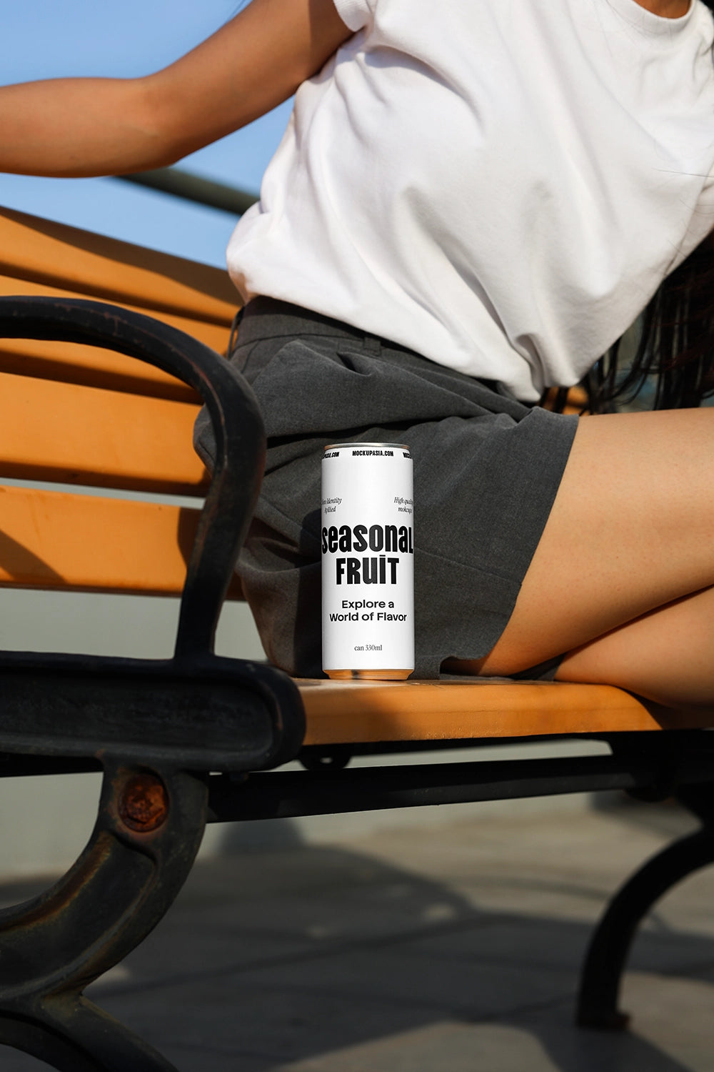 Soda can mockup placed on bench next to seated person with natural sunlight and minimal lifestyle scene