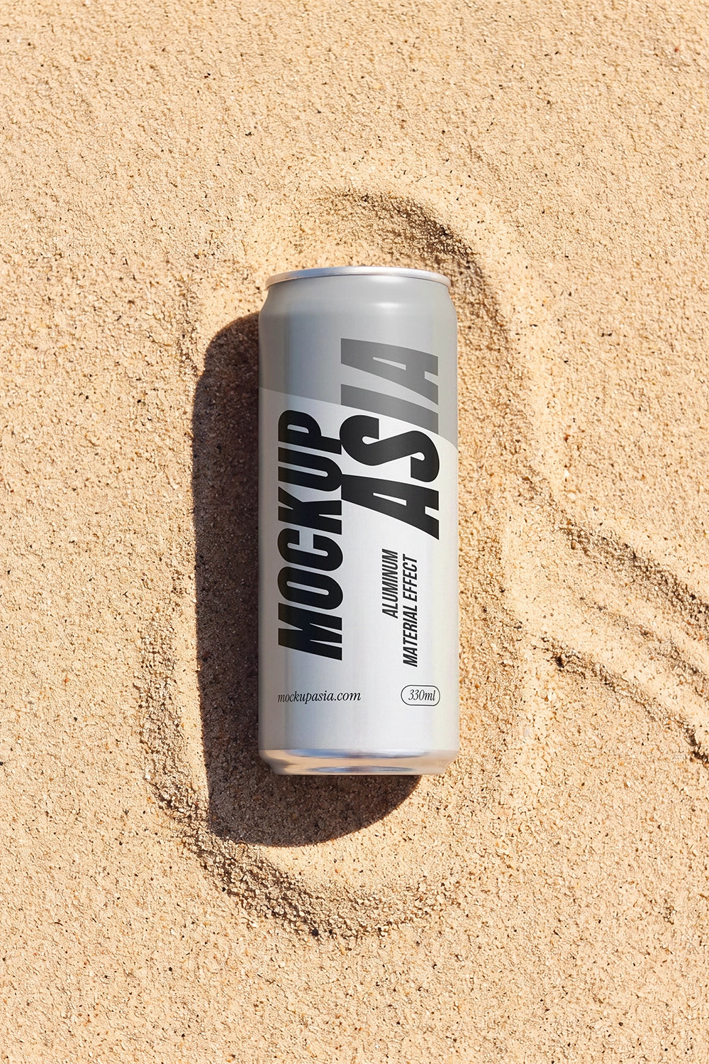 A silver aluminum beverage can with black text reading 'MOCKUP ASIA' lies in sandy beige beach sand, creating a small ring of sand around it.