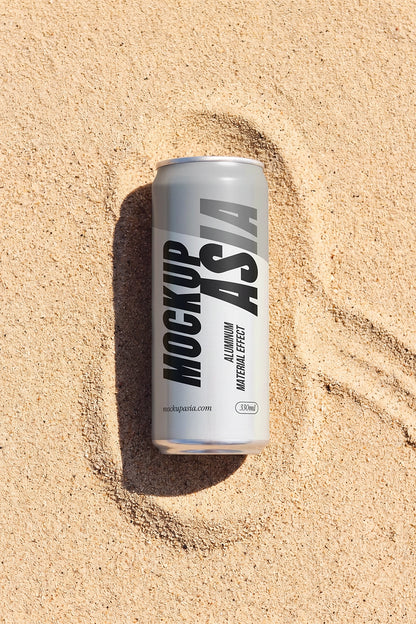 A silver aluminum beverage can with black text reading 'MOCKUP ASIA' lies in sandy beige beach sand, creating a small ring of sand around it.