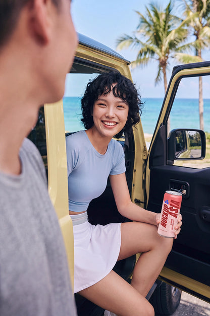A woman sitting in the open door of a yellow vehicle by the beach holds a pink can of Coca-Cola. A man in a gray shirt is partially visible in the foreground, with palm trees and blue ocean in the background.