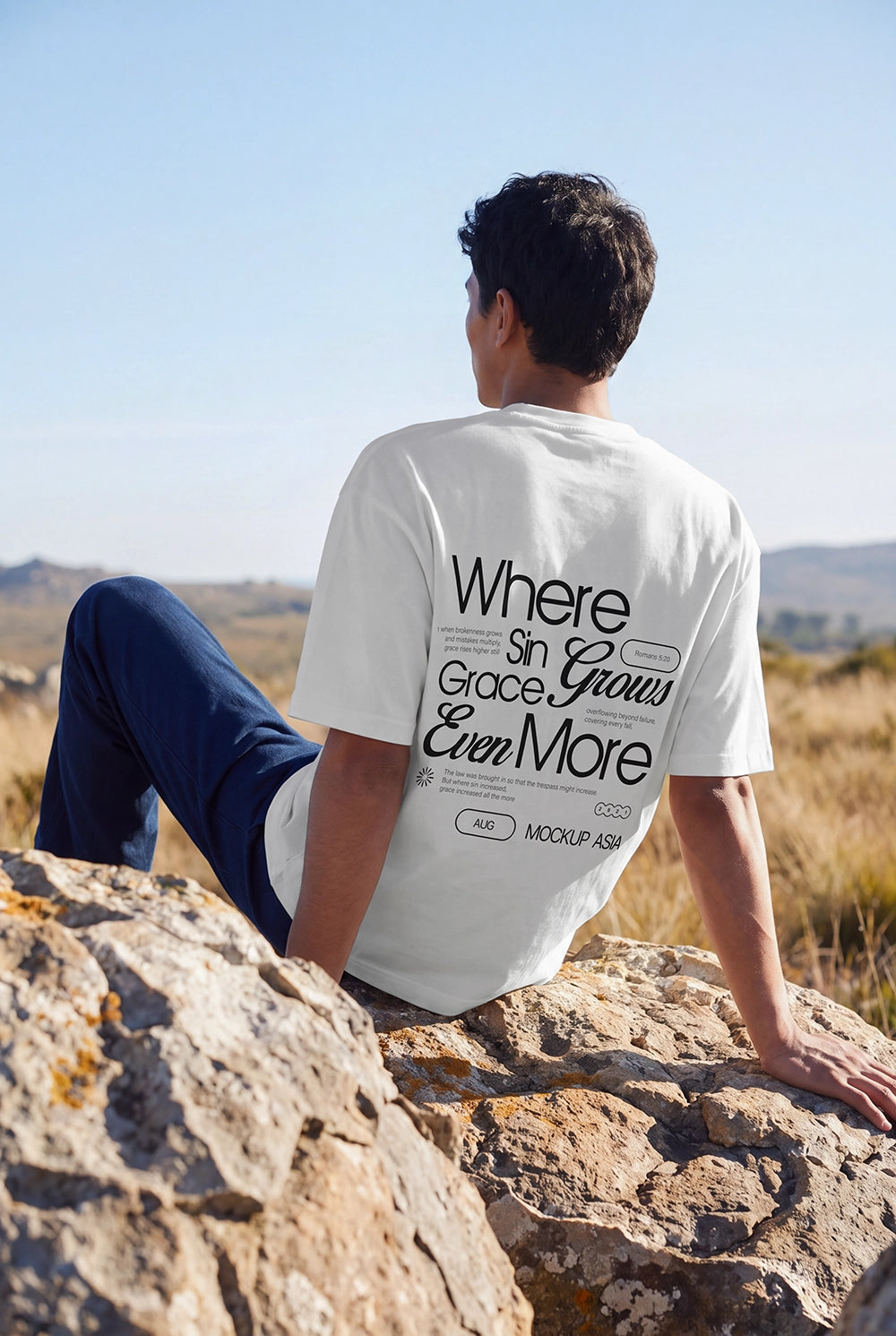 Man wearing an oversized white T-shirt with a custom back print, sitting on sunlit rocks in a natural outdoor landscape, apparel mockup.