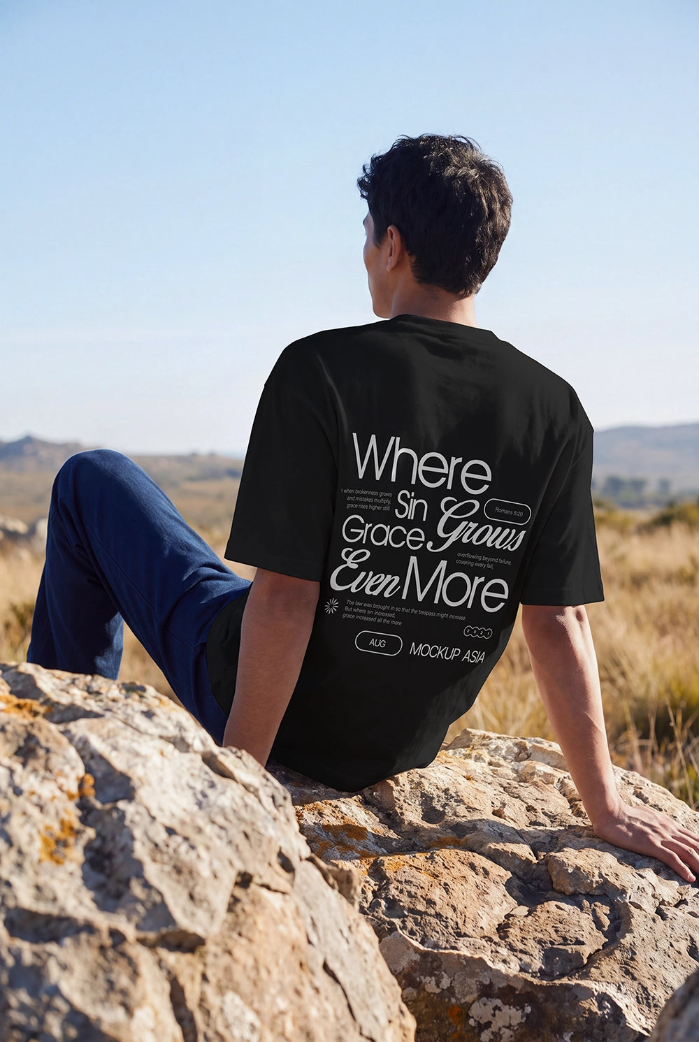 A man wearing a black graphic t-shirt and blue jeans sits on a large rock outdoors, looking up at a scenic landscape under a clear blue sky. The back of the t-shirt features a large white typographic design that reads 'Where Sin Grace Grows Ever More'.