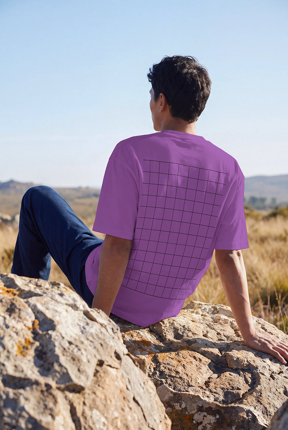A man sitting on a large rock outdoors, viewed from behind, wearing a purple short-sleeve t-shirt with a black grid pattern on the back and dark blue jeans.