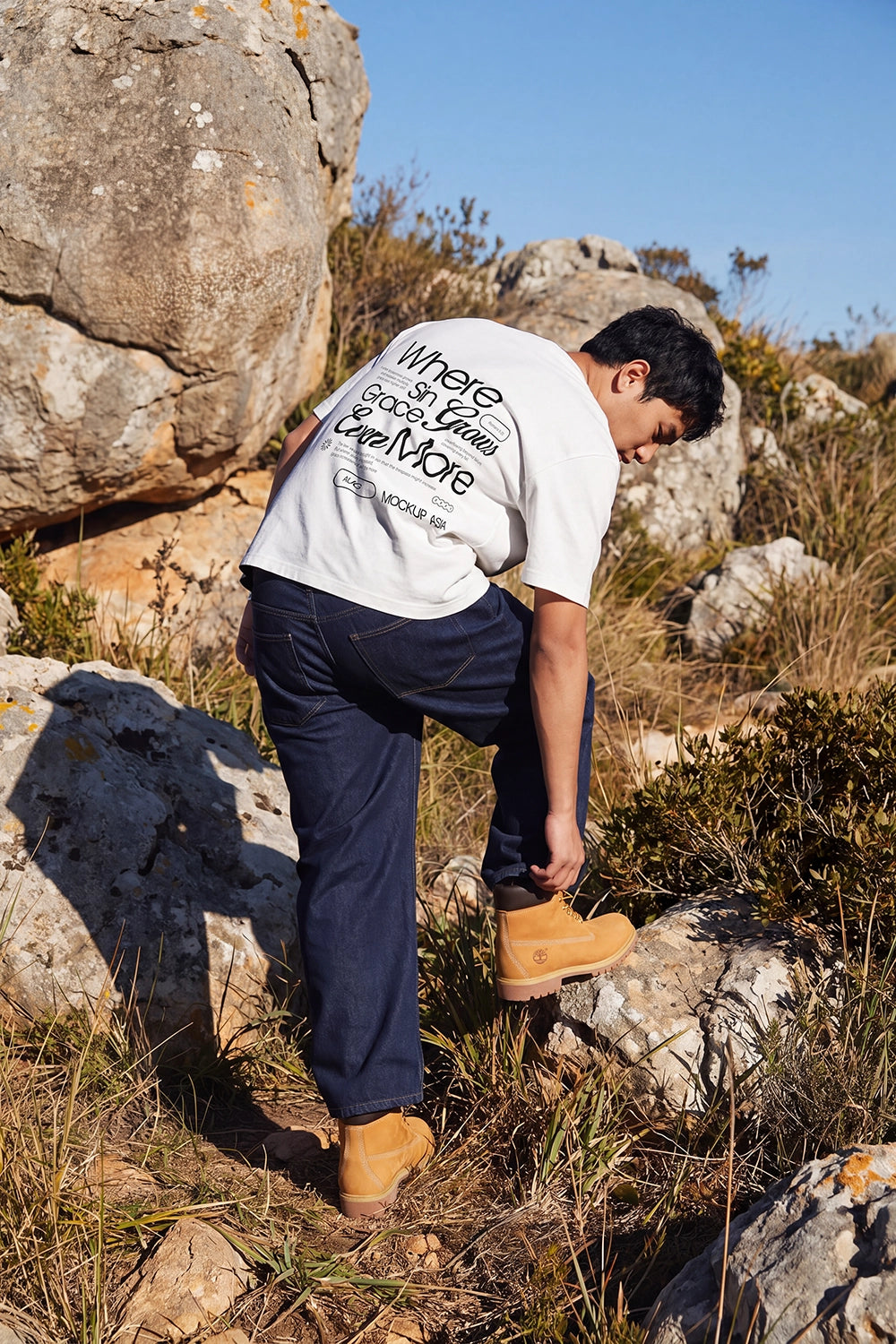 Man wearing an oversized white T-shirt with a custom back print while standing among sunlit rocks in a natural outdoor landscape.