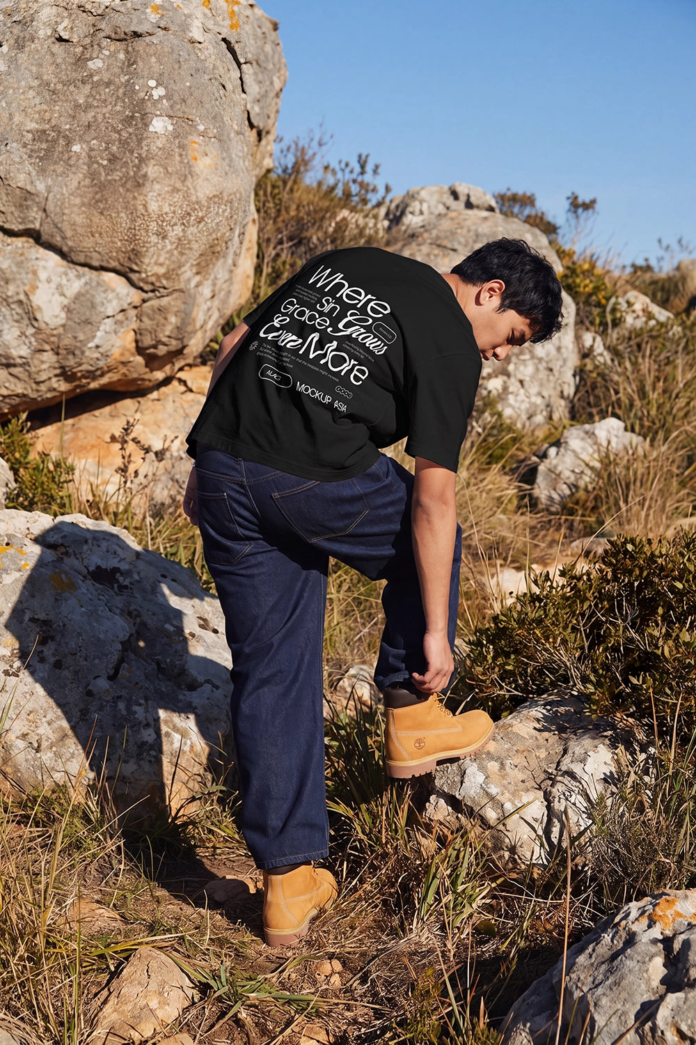 A man wearing a black graphic t-shirt, dark blue jeans, and tan boots is seen from behind while climbing rocks outdoors in a sunny, rocky landscape.