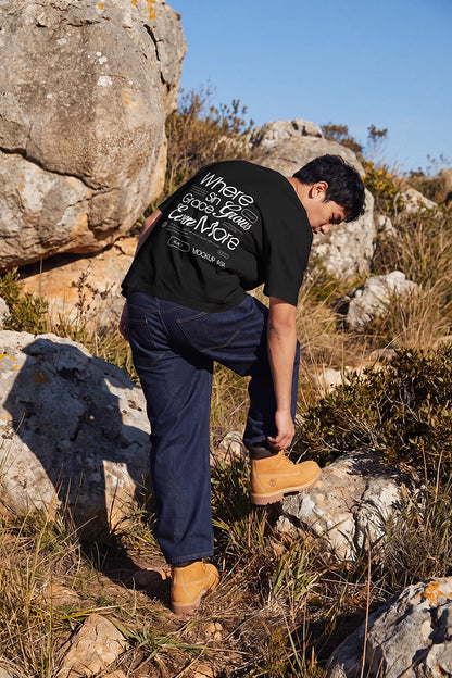 A man wearing a black graphic t-shirt, dark blue jeans, and tan boots is seen from behind while climbing rocks outdoors in a sunny, rocky landscape.