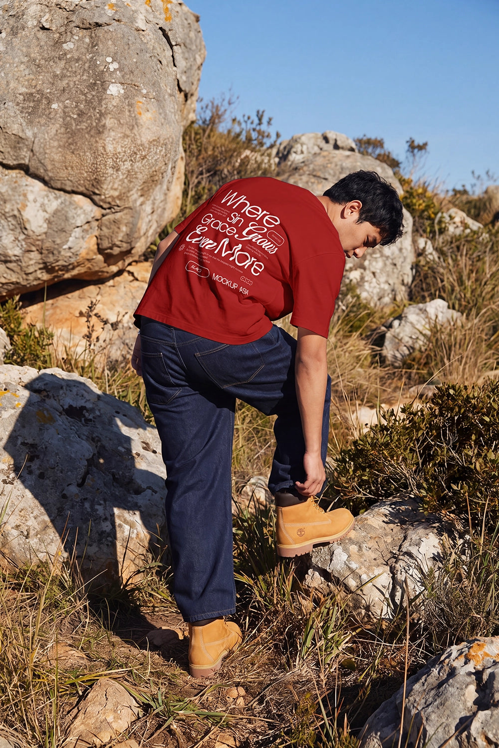 A man wearing a red graphic t-shirt, navy blue jeans, and tan boots is bent over on a rocky, grassy hillside outdoors.