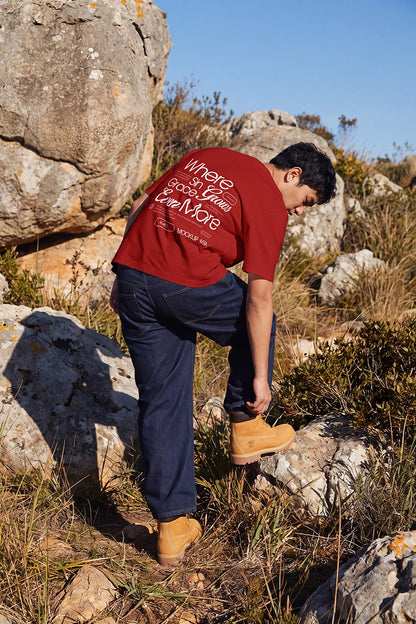 A man wearing a red graphic t-shirt, navy blue jeans, and tan boots is bent over on a rocky, grassy hillside outdoors.