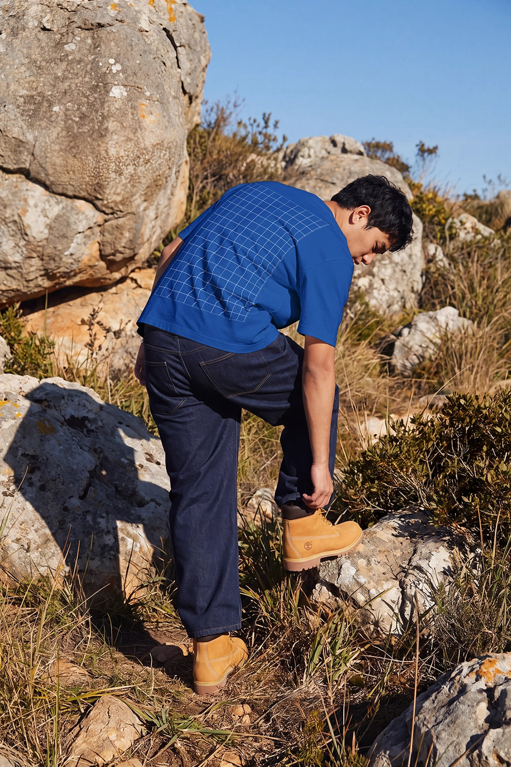 A man wearing a blue plaid short-sleeve shirt, navy trousers, and tan boots is bent over while climbing rocky, grassy terrain outdoors under a clear blue sky.