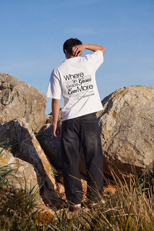 Man wearing a white oversized T-shirt with a custom back print standing between sunlit rocks in an outdoor landscape.