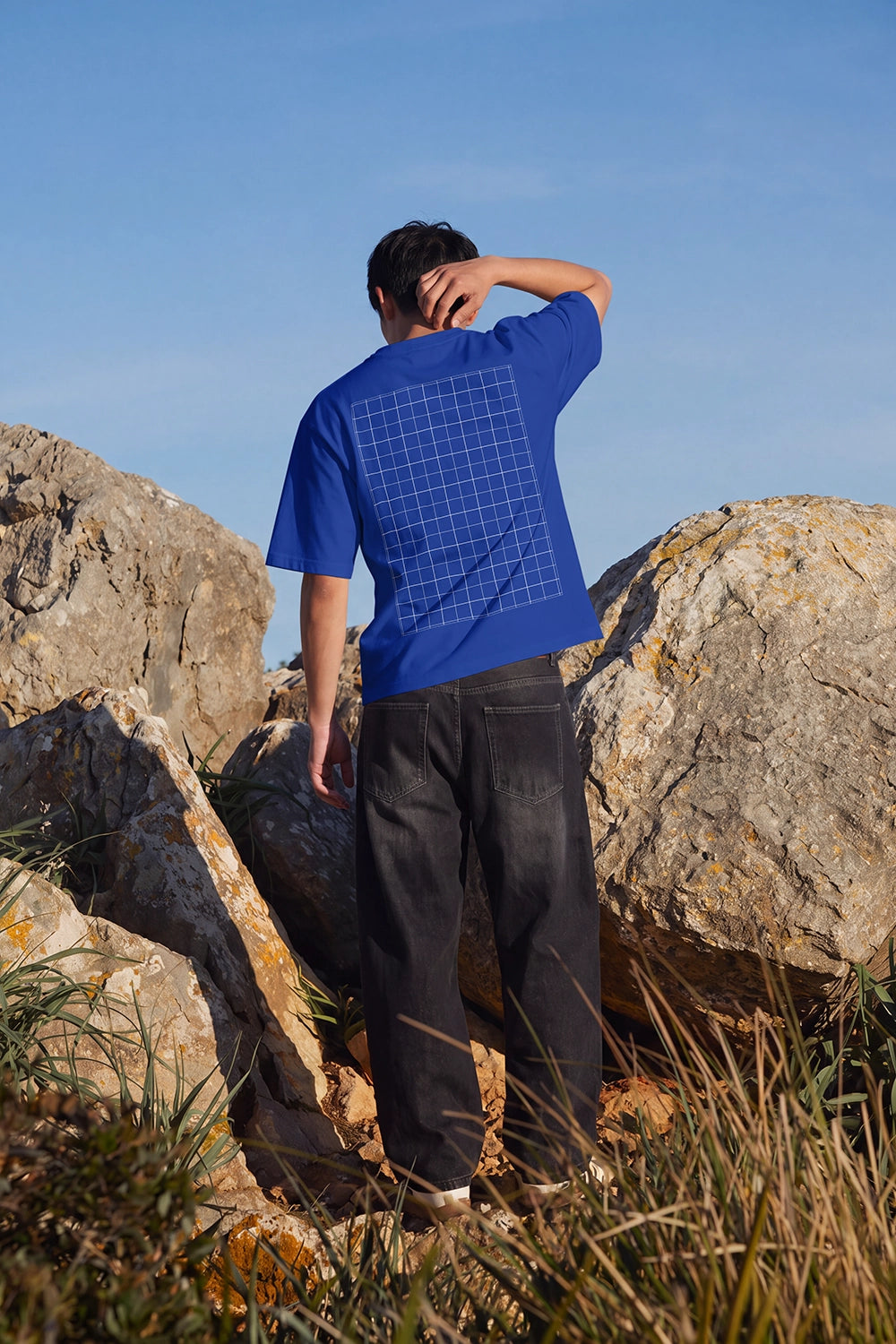 Full-body back view of a model wearing a custom graphic T-shirt surrounded by rugged rocks and natural scenery.