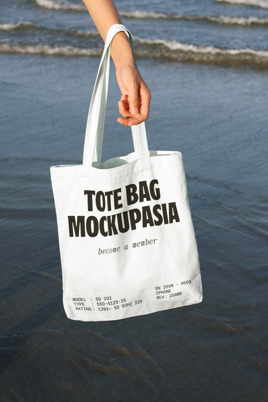 tote bag mockup held in hand at beach with water background and natural sunlight