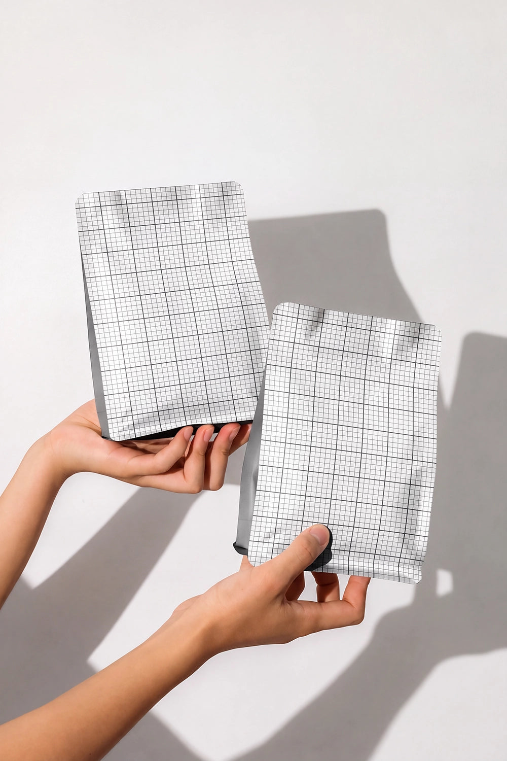 Two hands holding two small rectangular pouches with a white and gray grid pattern, set against a plain light gray background with soft shadows.