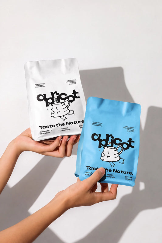 Two hands holding two bags of 'ourcot' cat food against a clean white background. One bag is white with black text and illustration, and the other is light blue with black text and illustration.
